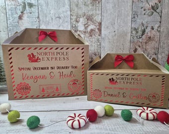 May include: Two brown cardboard gift boxes with red accents and bows. The boxes are labeled "North Pole Express" with custom names and "Special December 1st Delivery" text. A garland of colorful felt balls and candy canes decorates the scene.