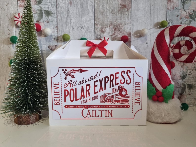 May include: A white gift box with a red bow on top. The box features a red graphic with the text "All aboard! Polar Express Train Ride" and the name "Caitlin". A small Christmas tree and a candy cane decoration are in the background.