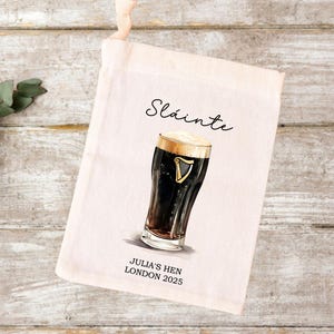 May include: A cream-colored tote bag featuring a watercolor illustration of a pint of dark beer with a foamy head. The word "Sláinte" is written above the beer, and below it reads "JULIA'S HEN LONDON 2025".