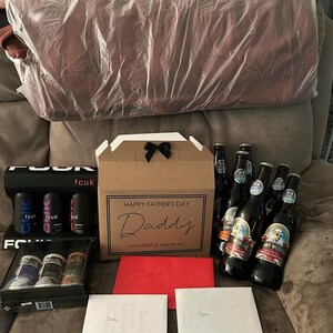 May include: A brown cardboard box with a black ribbon and the text "HAPPY FATHER'S DAY Daddy Love Eden & Harper xxx". The box is surrounded by several bottles of beer, some with labels that say "Henry Weston's Cider".