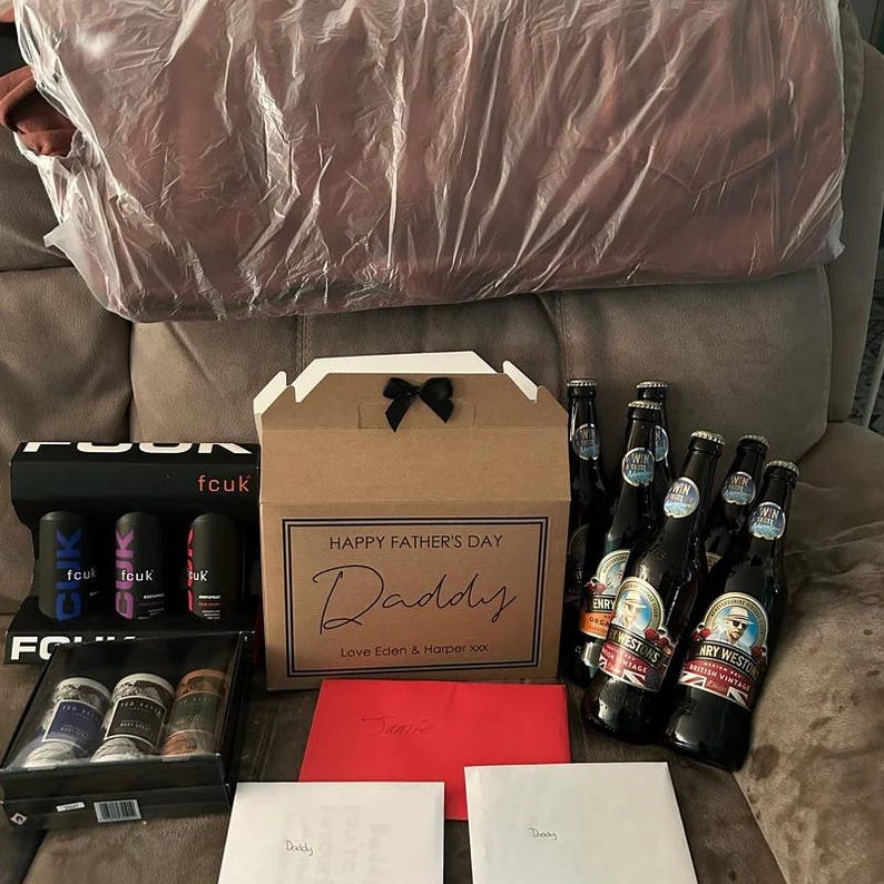 May include: A brown cardboard box with a black ribbon and the text "HAPPY FATHER'S DAY Daddy Love Eden & Harper xxx". The box is surrounded by several bottles of beer, some with labels that say "Henry Weston's Cider".