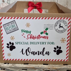 Puede incluir: Una caja de regalo de cart&oacute;n marr&oacute;n con un lazo rojo. La parte delantera tiene una etiqueta blanca con gr&aacute;ficos navide&ntilde;os, incluyendo huellas de patas, copos de nieve y el texto "Merry Christmas, Special Delivery For: Wanda".