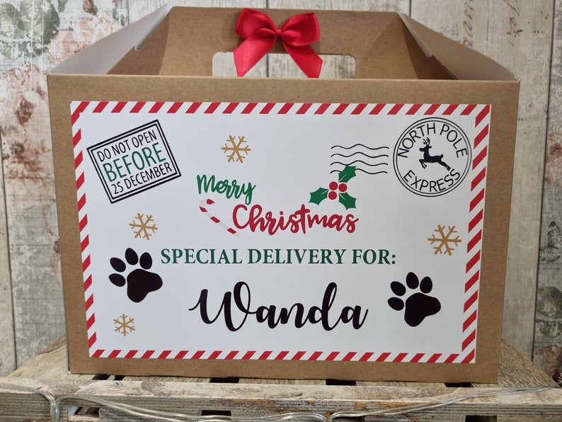 Puede incluir: Una caja de regalo de cart&oacute;n marr&oacute;n con un lazo rojo. La parte delantera tiene una etiqueta blanca con gr&aacute;ficos navide&ntilde;os, incluyendo huellas de patas, copos de nieve y el texto "Merry Christmas, Special Delivery For: Wanda".