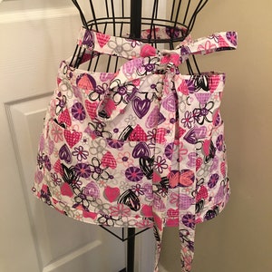 May include: A white apron with a pink, purple, and black heart and flower pattern. The apron has two pockets and ties at the waist.