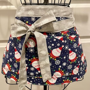 May include: A blue and white Christmas apron with a Santa Claus print and a gray snowflake patterned waistband. The apron has a pocket on the front.