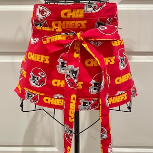 May include: Red apron with a repeating pattern of football helmets and the word "CHIEFS" in white. The apron has a yellow bow and long ties. The apron is displayed on a black metal stand.