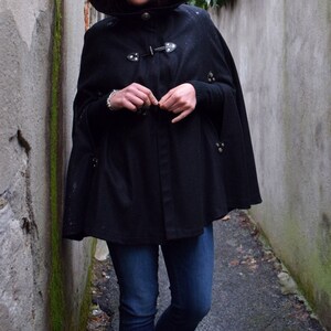 May include: A woman wearing a black hooded cape with a button closure and silver accents. She is wearing blue jeans and black boots.