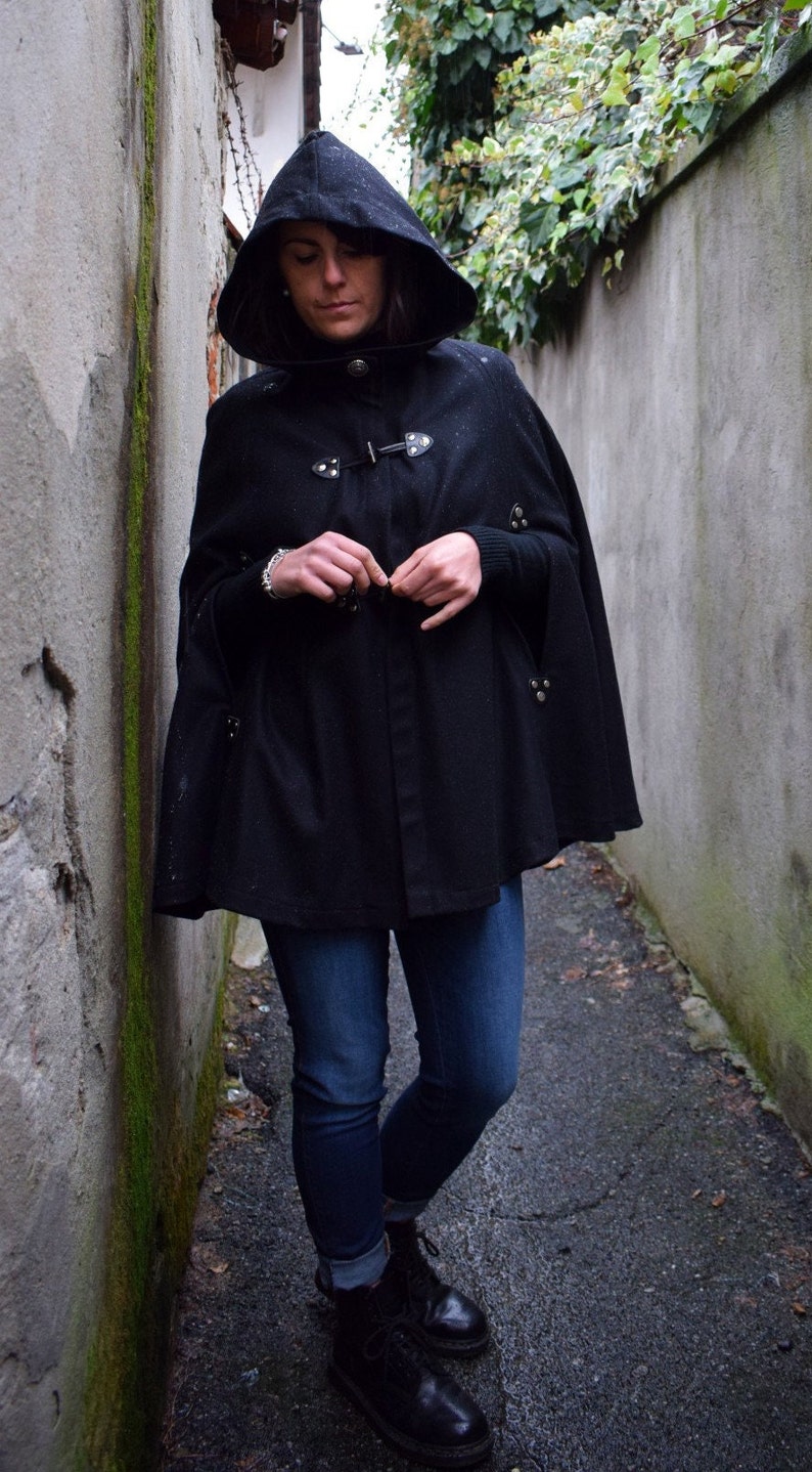 May include: A woman wearing a black hooded cape with a button closure and silver accents. She is wearing blue jeans and black boots.