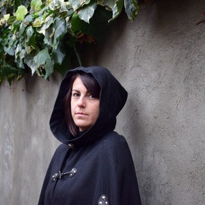 May include: A woman wearing a black hooded cloak with a high collar and a button fastening. The cloak has a textured fabric and is lined with a soft material. The woman is standing in front of a grey wall with ivy growing on it.