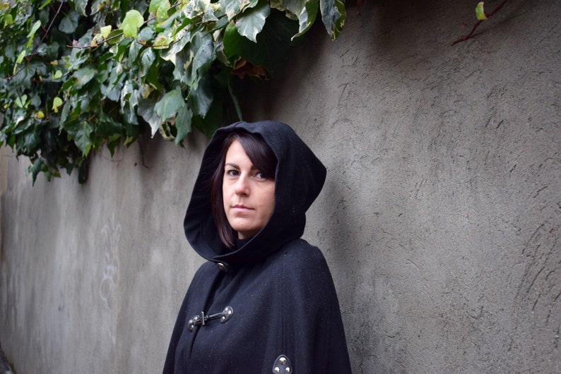 May include: A woman wearing a black hooded cloak with a high collar and a button fastening. The cloak has a textured fabric and is lined with a soft material. The woman is standing in front of a grey wall with ivy growing on it.