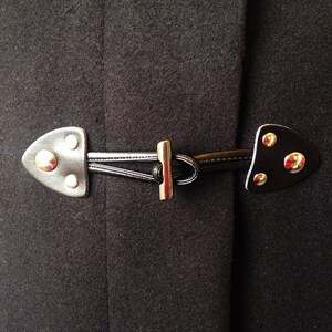 May include: A black wool coat with a silver metal toggle closure. The toggle is made of two pieces of leather with metal accents.