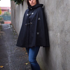 May include: A woman wearing a black hooded cape with silver accents and blue jeans. She is standing in a narrow alleyway with a concrete wall behind her.