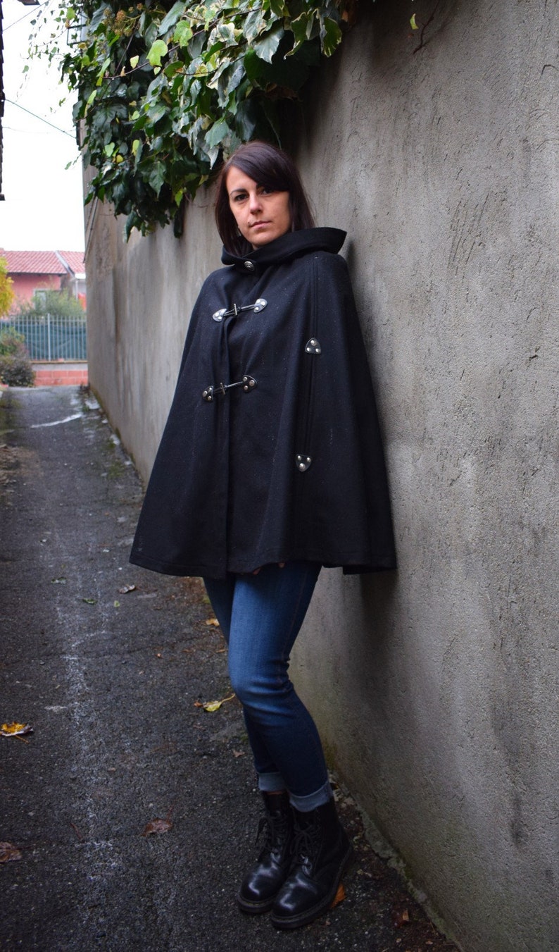 May include: A woman wearing a black hooded cape with silver accents and blue jeans. She is standing in a narrow alleyway with a concrete wall behind her.