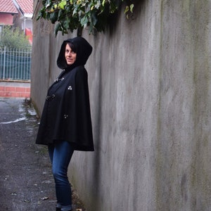 May include: A woman wearing a black hooded cape with silver accents and blue jeans. She is standing in front of a grey concrete wall.
