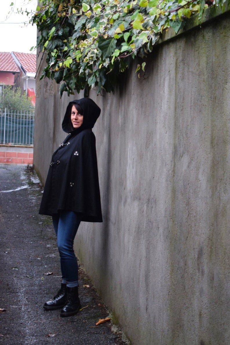 May include: A woman wearing a black hooded cape with silver accents and blue jeans. She is standing in front of a grey concrete wall.