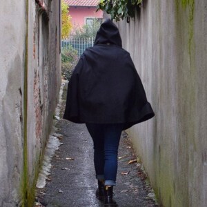 May include: A person wearing a black hooded cloak walks down a narrow alleyway between two grey brick walls.