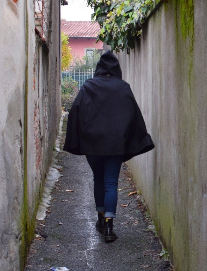 May include: A person wearing a black hooded cloak walks down a narrow alleyway between two grey brick walls.
