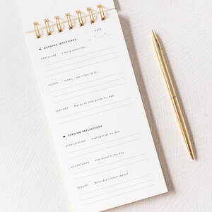 May include: A white journal with a gold spiral binding. The journal has a black and white design with the words "Morning Intentions" and "Evening Reflections" printed on the cover. The journal has sections for gratitude, vision, support, appreciation, acceptance, and inquiry.