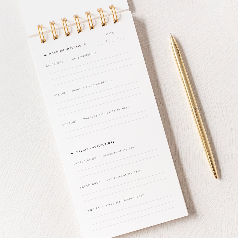 May include: A white journal with a gold spiral binding. The journal has a black and white design with the words "Morning Intentions" and "Evening Reflections" printed on the cover. The journal has sections for gratitude, vision, support, appreciation, acceptance, and inquiry.