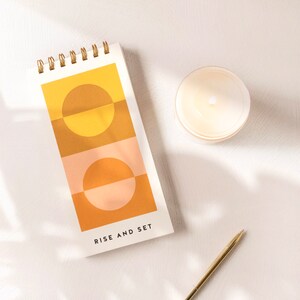 May include: A white notepad with a gold spiral binding. The notepad has a geometric design with two orange and yellow circles on a peach and orange background. The text "Rise and Set" is printed at the bottom of the notepad.