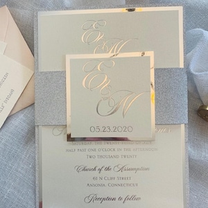 May include: A wedding invitation with a silver glitter background and a light blue inner card. The invitation features the date "05.23.2020", the text "Saturday, the twenty-third of May", "Half past one o'clock in the afternoon", "Two thousand twenty", "Church of the Assumption", "61 N Cliff Street", "Ansonia, Connecticut", and "Reception to follow".