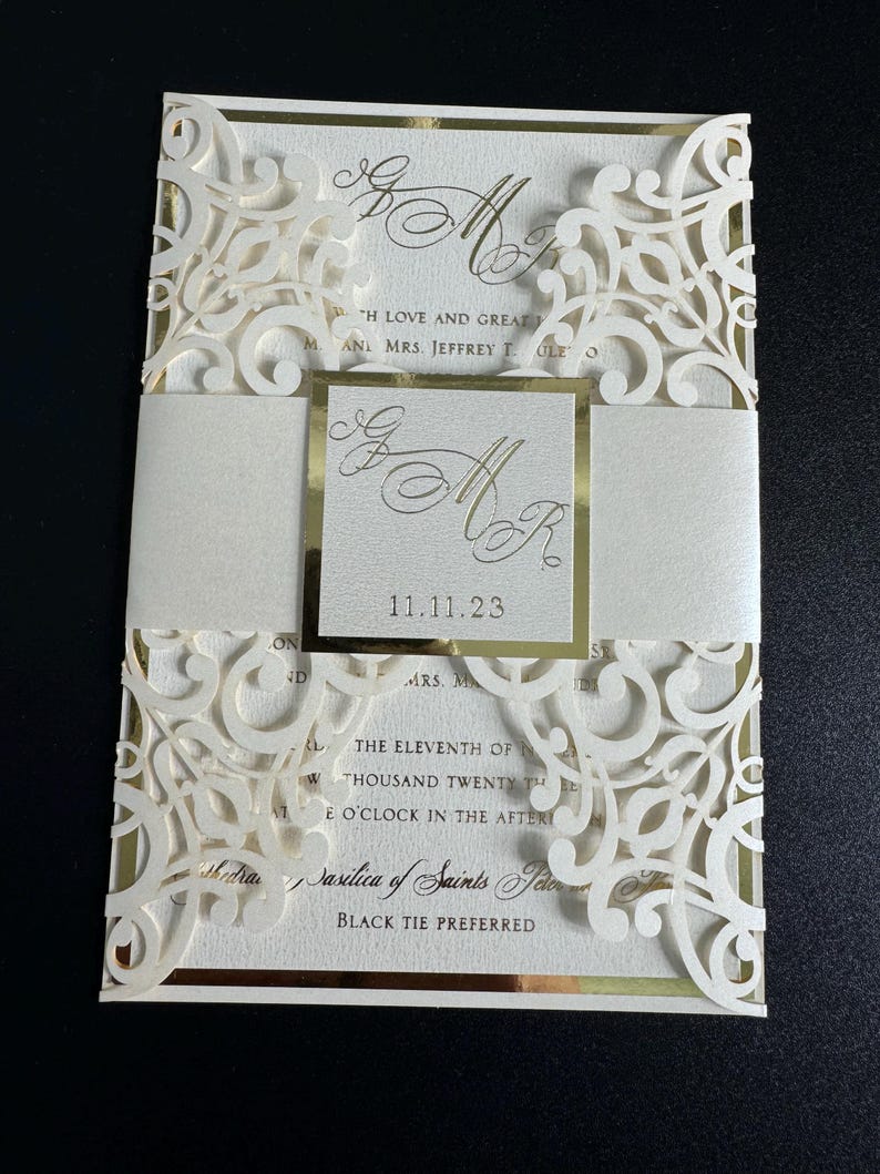 Op de afbeelding: Een witte huwelijksuitnodiging met gouden accenten en een lasergesneden ontwerp. De uitnodiging bevat de datum "11.11.23" en de tekst "Basilica of Saints Peter and Paul" en "Black Tie Preferred".
