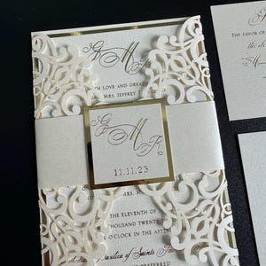 Op de afbeelding: Een witte huwelijksuitnodiging met gouden accenten en een lasergesneden ontwerp. De uitnodiging bevat de datum "11.11.23" en de tekst "Basilica of Saints Peter and Paul" met "Black Tie Preferred" eronder.