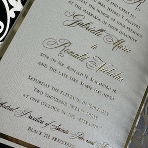 Op de afbeelding: Gouden huwelijksuitnodiging met een witte achtergrond en zwarte en gouden decoratieve accenten. De uitnodiging kondigt het huwelijk aan van Gabrielle Maric en Ronald Nicholas op zaterdag 11 november 2023 om 13:00 uur in de Basiliek van de Heiligen Petrus en Paulus. Zwarte stropdas is gewenst.