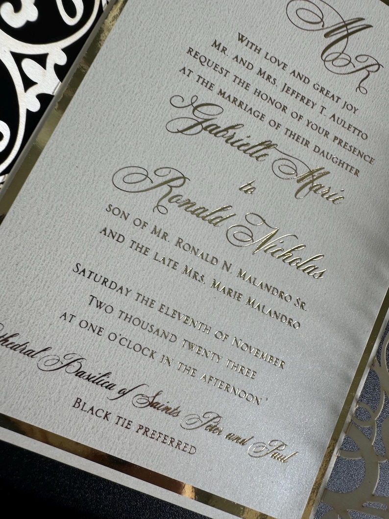 Op de afbeelding: Gouden huwelijksuitnodiging met een witte achtergrond en zwarte en gouden decoratieve accenten. De uitnodiging kondigt het huwelijk aan van Gabrielle Maric en Ronald Nicholas op zaterdag 11 november 2023 om 13:00 uur in de Basiliek van de Heiligen Petrus en Paulus. Zwarte stropdas is gewenst.