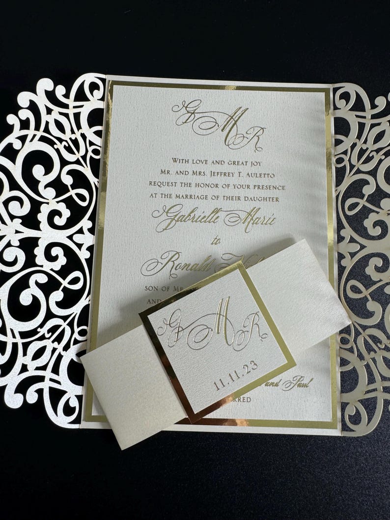 Op de afbeelding: Een witte en gouden huwelijksuitnodiging met een decoratief kantontwerp. De uitnodiging bevat de namen "Gabrielle Marie" en "Ronald" met de datum "11.11.23".