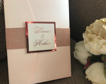 May include: A blush pink wedding invitation with a rose gold glitter band and a rose gold foil square with the text "Diana & Hakam" in cursive.