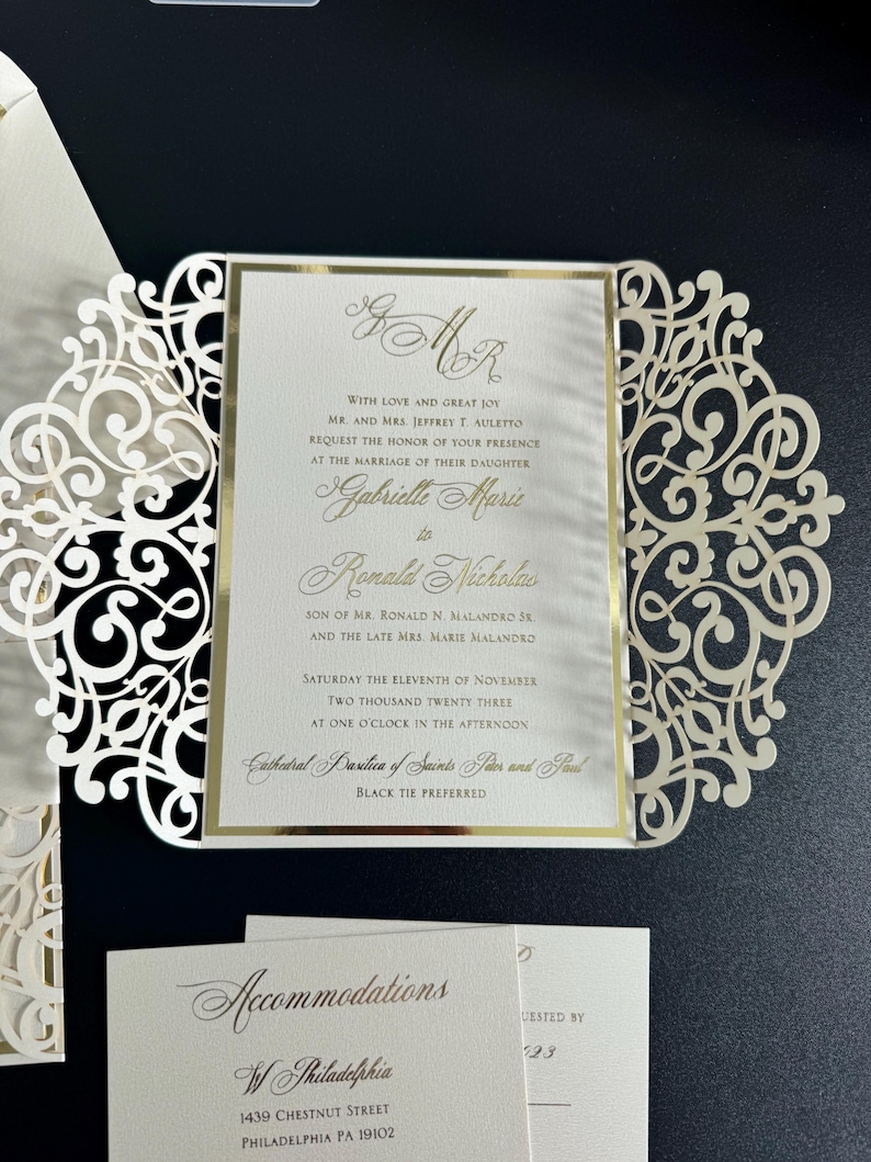 Op de afbeelding: Een witte en gouden huwelijksuitnodiging met een decoratief kantontwerp. De uitnodiging kondigt het huwelijk aan van Gabrielle Marie en Ronald Nicholas op zaterdag 11 november 2023 om 13:00 uur in de kathedraalbasiliek van de Heiligen Petrus en Paulus. Zwarte das is gewenst.