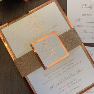 May include: A wedding invitation with a gold glitter background and a copper border. The invitation features the couple's initials, "S & H", in a cursive font. The text on the invitation reads "Sunday, the twenty sixth of May, two thousand and nineteen, at half past two o'clock in the afternoon, A Natalia Grand Celebration, 13 S Mt Vernon Ave, Atlantic City, New Jersey, Black Tie Optional".