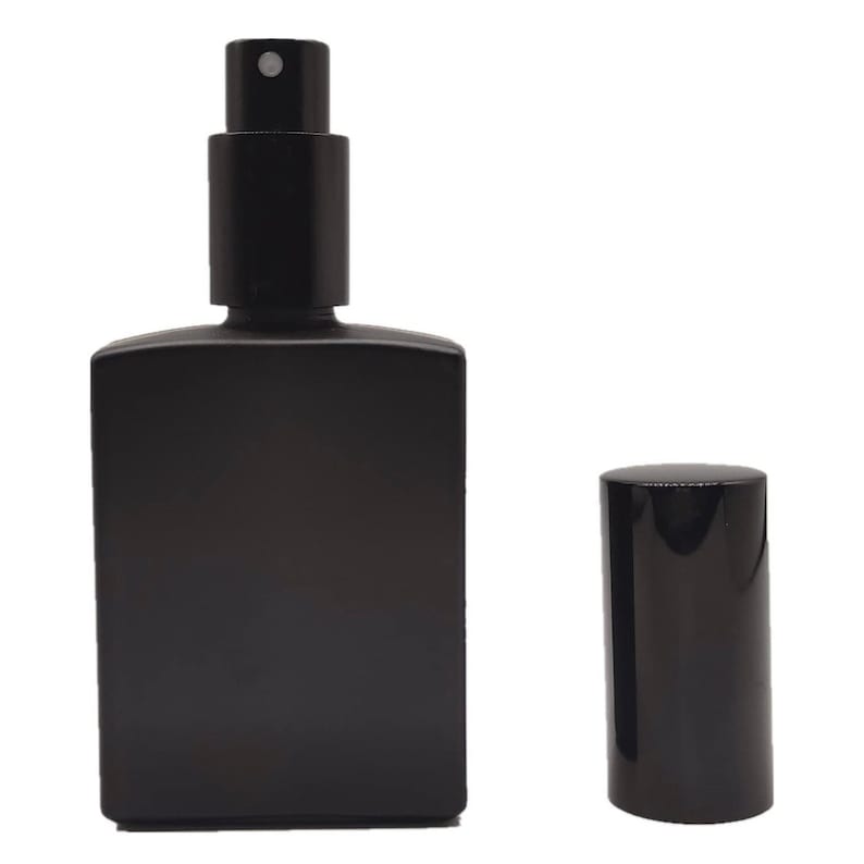 K&ouml;nnte beinhalten: Eine rechteckige Parf&uuml;mflasche aus mattem schwarzem Glas mit einem schwarzen Spr&uuml;hmechanismus und einer separaten zylindrischen schwarzen Kappe, die rechts daneben steht. Die Flasche hat eine glatte, undurchsichtige Oberfl&auml;che und einen Spr&uuml;hkopf.