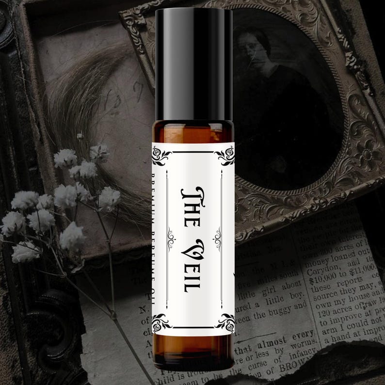 K&ouml;nnte beinhalten: Eine zylindrische Parf&uuml;m&ouml;l-Roll-on-Flasche aus bernsteinfarbenem Glas mit schwarzem Verschluss. Das wei&szlig;e rechteckige Etikett zeigt schwarze dekorative Schn&ouml;rkelr&auml;nder und den Text THE VEIL in fetter Serifenschrift sowie PREMIUM PERFUME OIL.