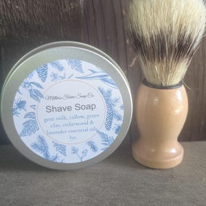May include: A round, silver tin of shave soap with a blue and white label, next to a shaving brush with a light brown handle and white bristles. The label reads "Shave Soap" and lists ingredients.
