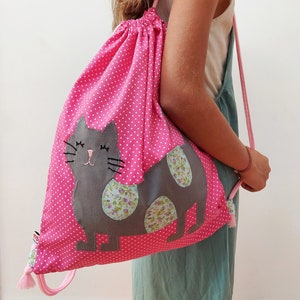 May include: Pink drawstring bag with white polka dots and a gray cat appliqué with floral accents. The cat has closed eyes and a pink nose.