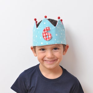 Può includere: Un bambino piccolo che indossa una corona blu e bianca con stelle rosse e un numero sei a scacchi rossi e bianchi. La corona ha dei pompon rossi in cima.