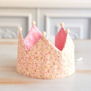 May include: A pink and white fabric crown with a floral pattern and gingham trim. The crown has white pom-poms around the edge.