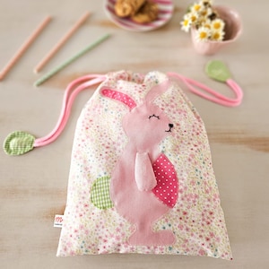 Può includere: Una borsa a cordoncino rosa con un'applicazione a forma di coniglio. Il coniglio ha orecchie rosa, un corpo rosa e una coda a scacchi verde e bianca. La borsa è realizzata in tessuto bianco con un motivo floreale rosa.