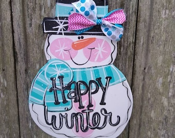 May include: A white snowman door hanger with a black top hat, a pink and blue bow, and the words "Happy Winter" in black script.