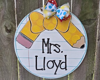 May include: A round, wooden teacher's sign with a pencil design and the name "Mrs. Lloyd" in black lettering. The sign has a blue border and is topped with a colorful bow.