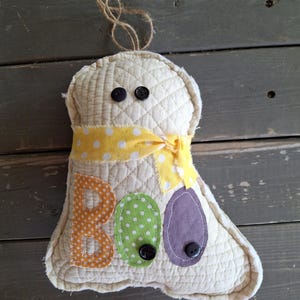 May include: A quilted ghost decoration with the word "BOO" stitched on the front. The ghost is cream-colored with a yellow polka-dot ribbon and black button eyes. The letters are orange, green, and purple.