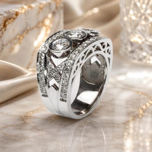 A silver ring with a large central diamond surrounded by smaller diamonds, set in a decorative band, is displayed on a marble surface.