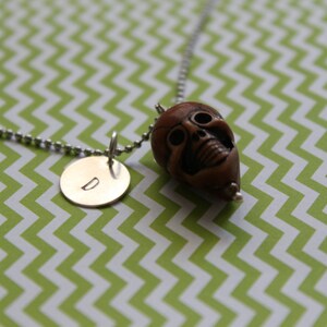 May include: A silver ball chain necklace with two pendants. One pendant is a small, brown skull. The other is a gold-colored, circular pendant with the letter "D" engraved on it. The background is a green and white chevron pattern.