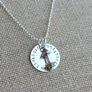 May include: A silver necklace with a round pendant featuring a dumbbell charm. The pendant has the words "progress not perfection" engraved around the edge. The necklace is displayed on a textured, tan surface.