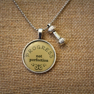 May include: Silver-toned necklace with a round pendant that reads "PROGRESS not perfection" in black text. A small dumbbell charm hangs next to the pendant. The necklace is displayed on a textured, tan surface.