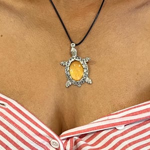 May include: A silver-toned turtle pendant necklace with an oval, amber-colored stone center. The pendant is suspended from a black cord. The person is wearing a red and white striped shirt.