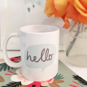Op de afbeelding: Witte keramische koffiemok met een lichtblauwe spraakballon waarop in grijs schrift "hello" staat. De mok staat op een tafel met een bloemenpatroon.