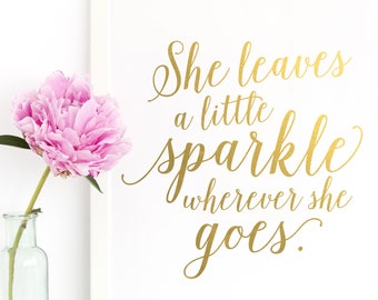 Puede incluir: Una impresión enmarcada con una escritura dorada que dice: "She leaves a little sparkle wherever she goes". Una peonía rosa está en un jarrón de vidrio transparente a la izquierda de la impresión. La impresión está en un marco blanco.